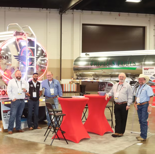 Five men standing and smiling near two red-covered tables at an indoor trade show booth with large fuel tanks and trailers displayed behind them.